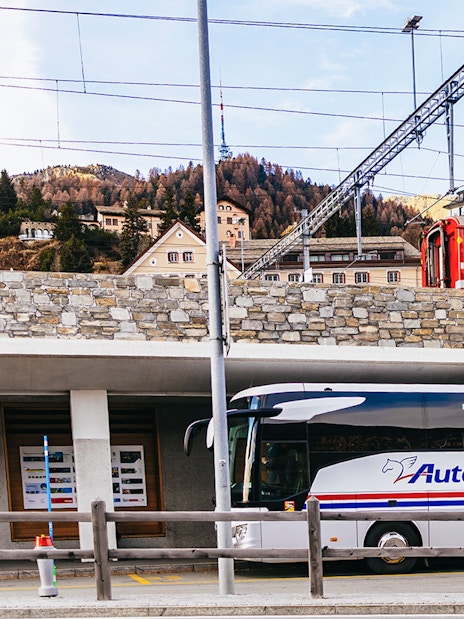 Bernina Red Train passing through St. Moritz station with a tour bus parked below.