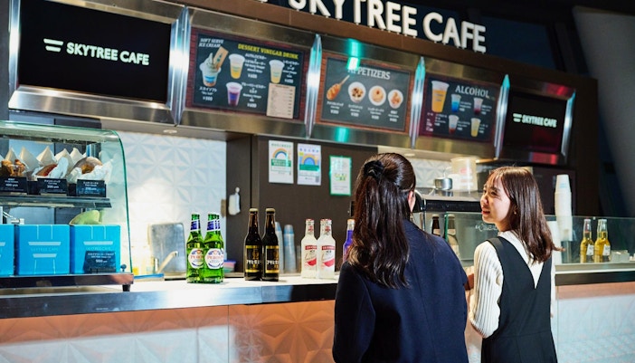 Tourists enjoying drinks at Skytree Cafe on Tokyo's Tembo Deck with city skyline views.