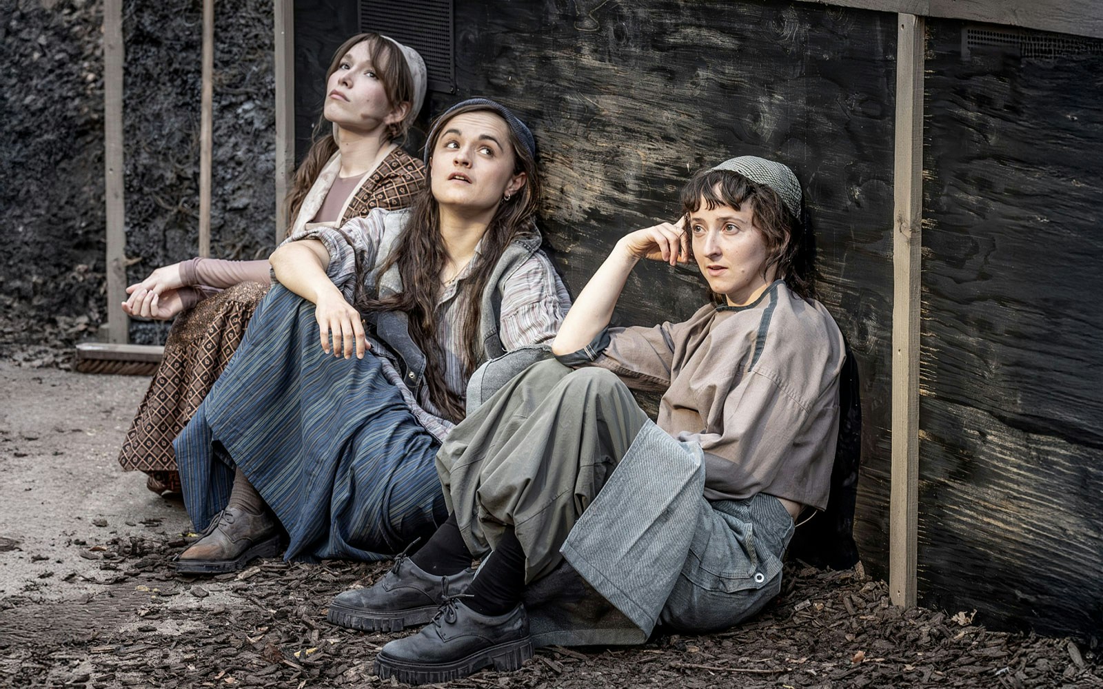 Three actors in period costumes sitting against a wooden wall, scene from Fiddler on the Roof.