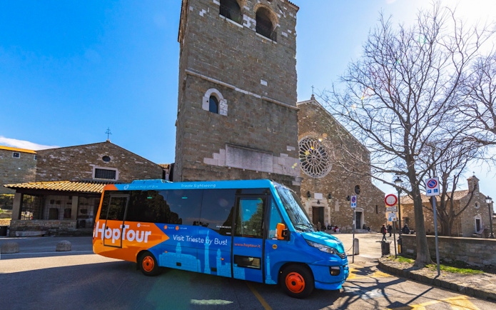 Hop-on hop-off bus in front of historic church in Trieste, Italy.