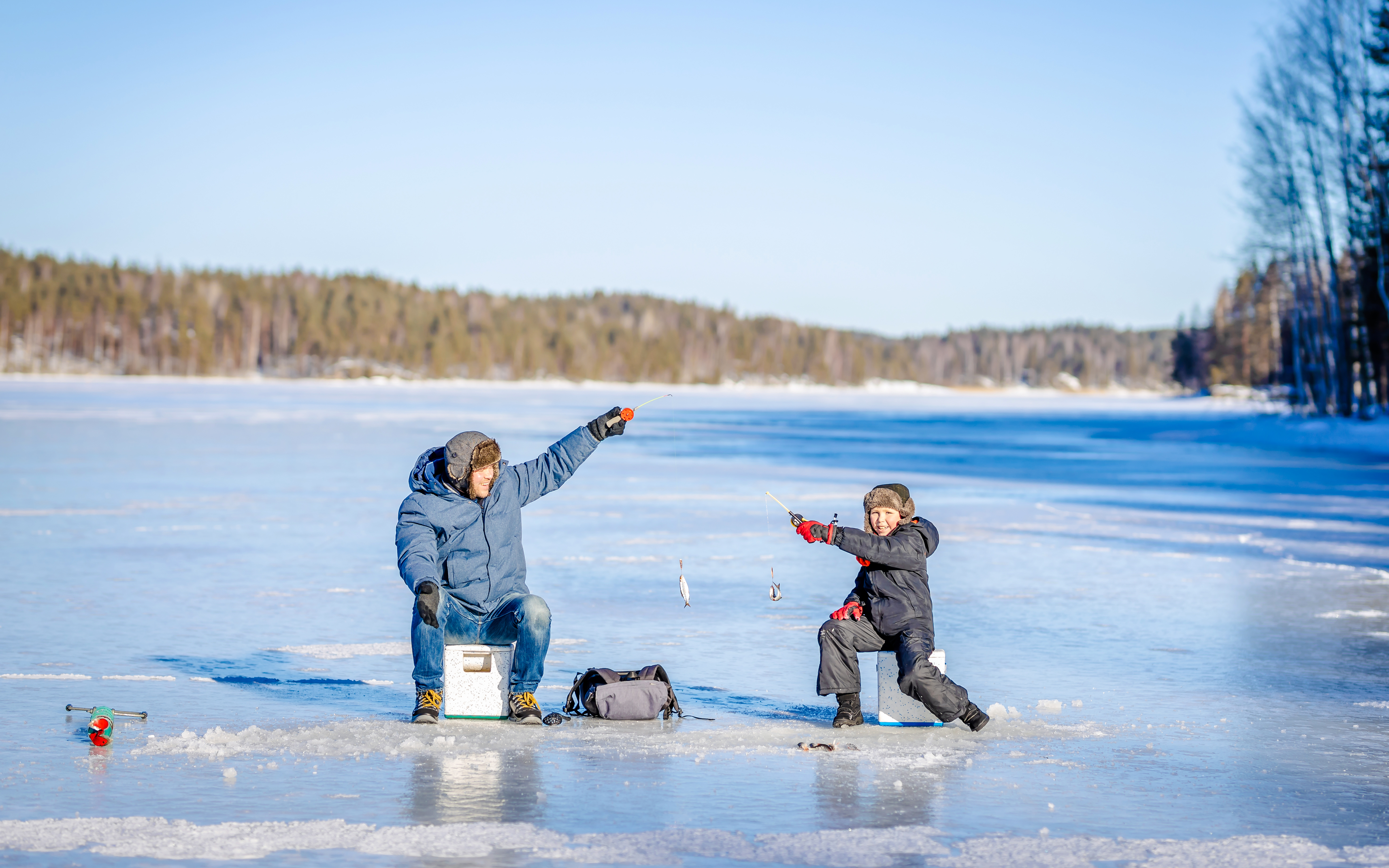 Father and son ice fishing on a frozen lake with forest in the background.