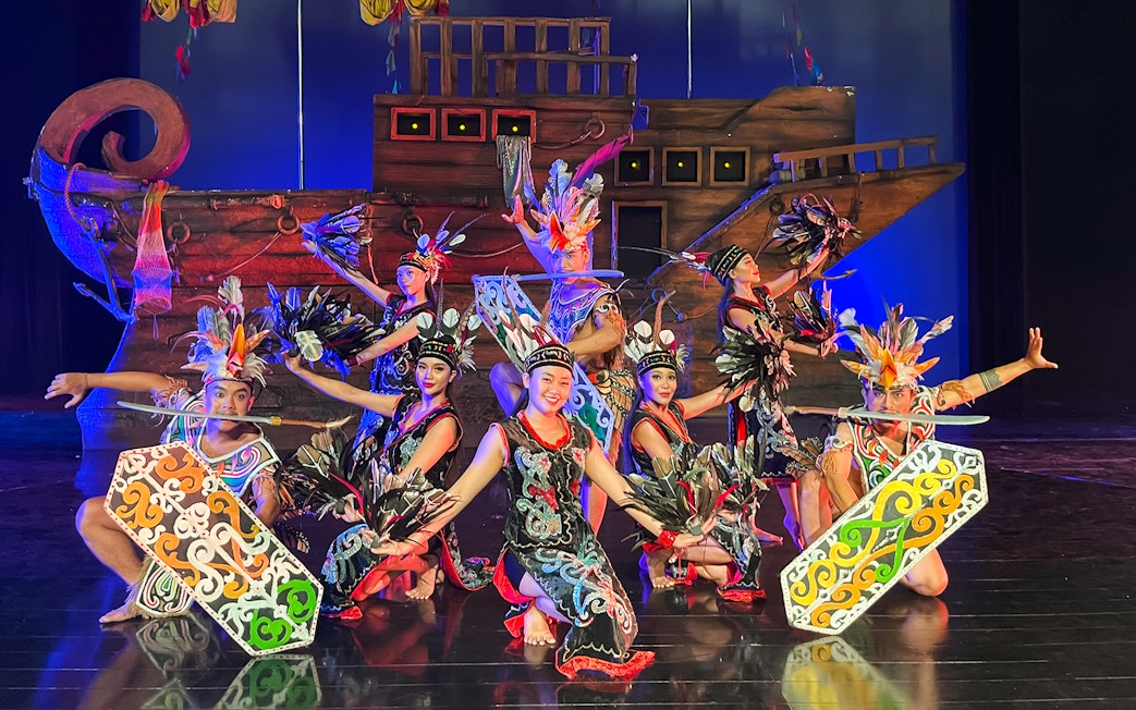 Performers in traditional Indonesian costumes at the Devdan Show, Bali, with a ship backdrop.