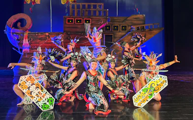 Performers in traditional Indonesian costumes at the Devdan Show, Bali, with a ship backdrop.