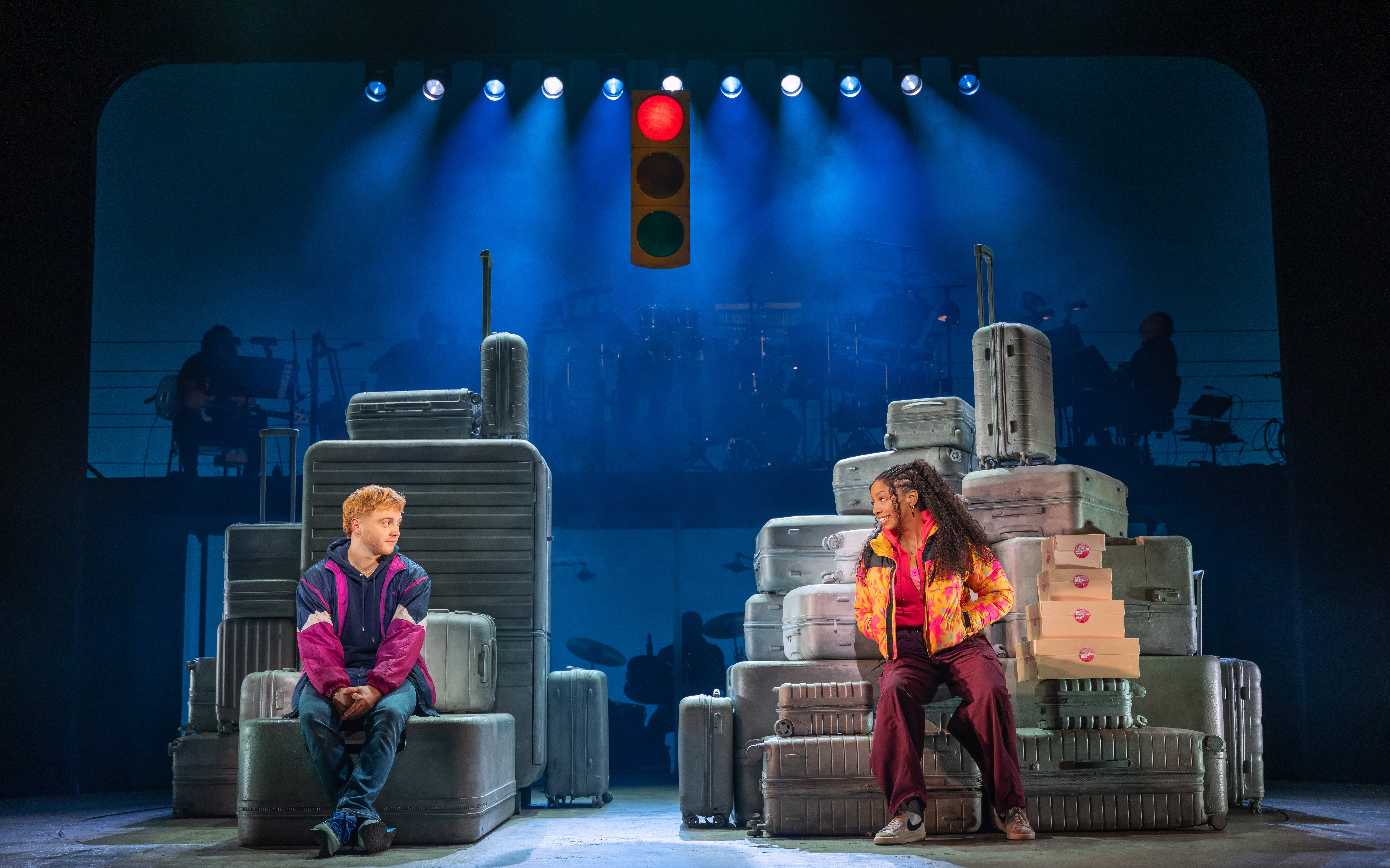 Two actors sit on stacked suitcases under a traffic light in the Broadway show "Two Strangers (Carry a Cake Across New York).