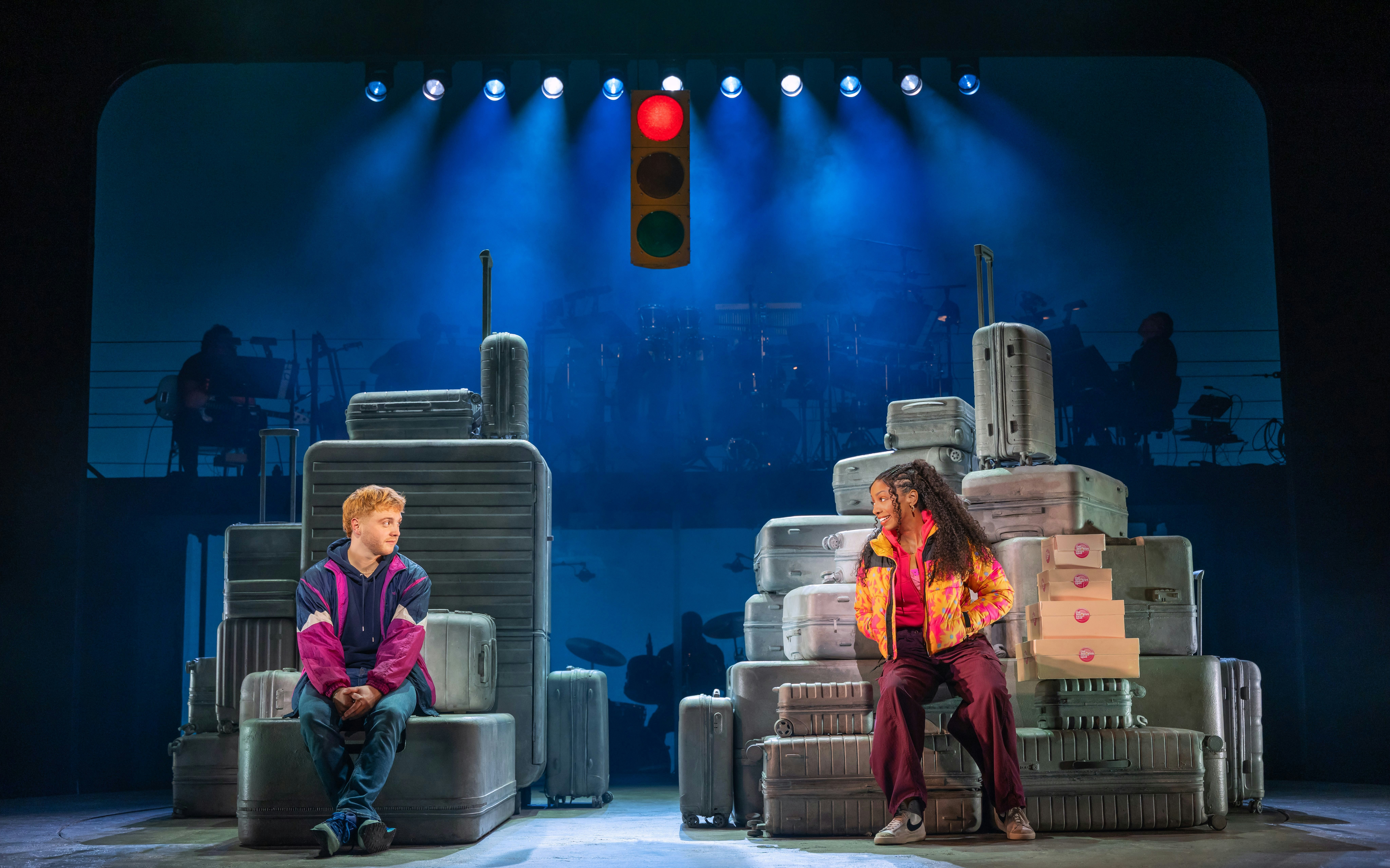 Two actors sit on stacked suitcases under a traffic light in the Broadway show "Two Strangers (Carry a Cake Across New York).