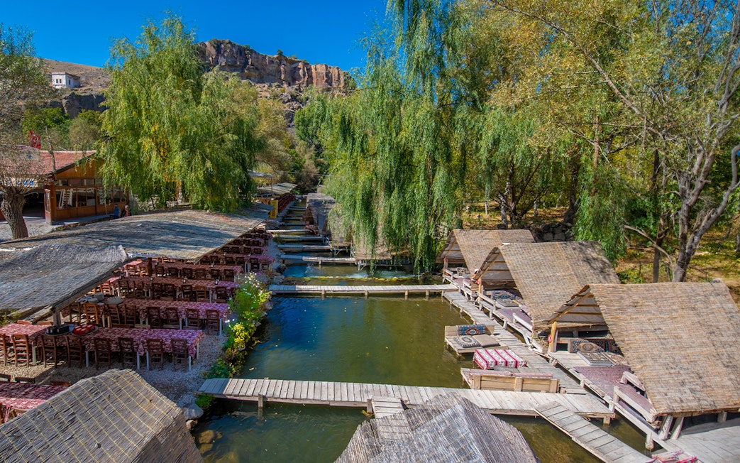 Outdoor dining area with wooden huts and tables by a river in Cappadocia, Turkey.