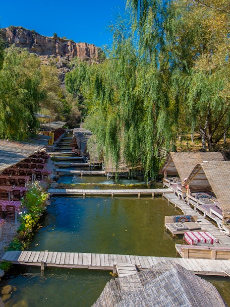 Outdoor dining area with wooden huts and tables by a river in Cappadocia, Turkey.