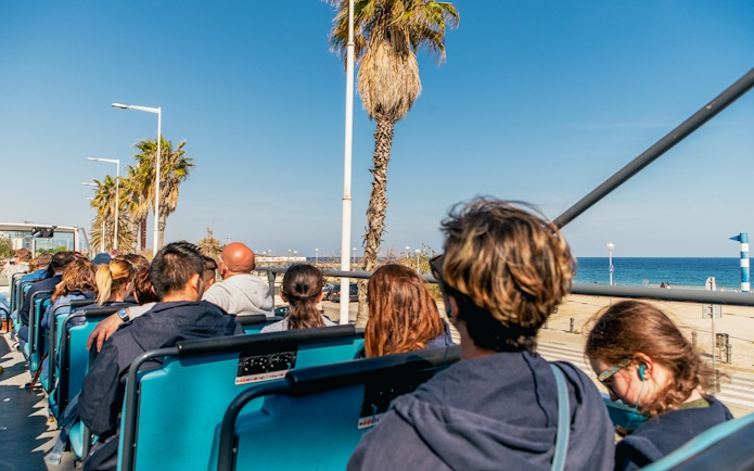 Tourists on a Barcelona hop-on hop-off bus with a view of the beach and cityscape.