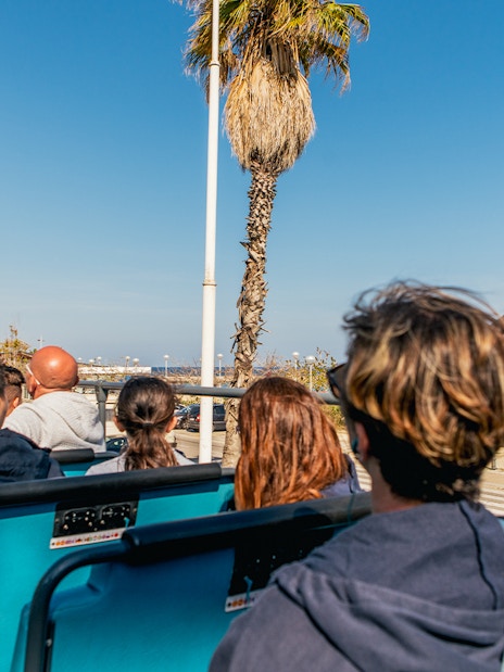 Tourists on a Barcelona hop-on hop-off bus with a view of the beach and cityscape.
