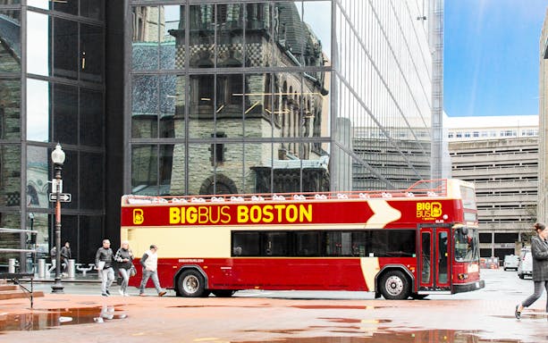 Open-top tour bus in Boston with city architecture reflected in glass building.