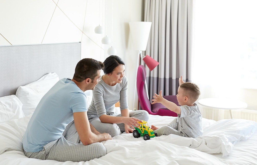 Family relaxing in a hotel room with a city view, featuring parents and child.
