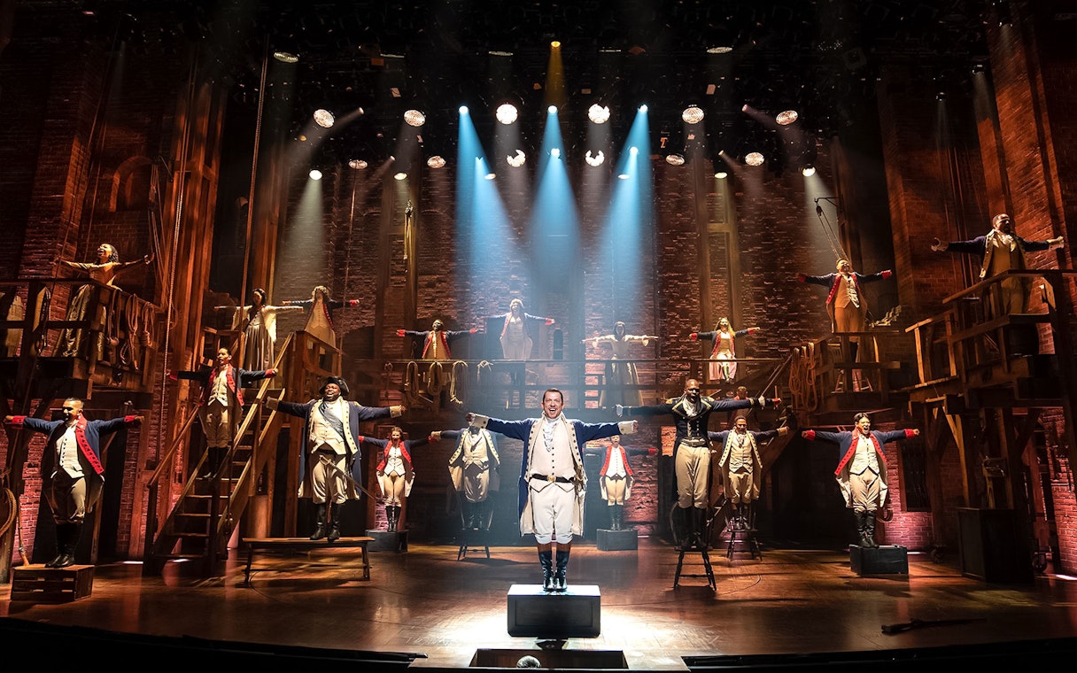 Stage performance of Hamilton cast in period costumes with dramatic lighting.