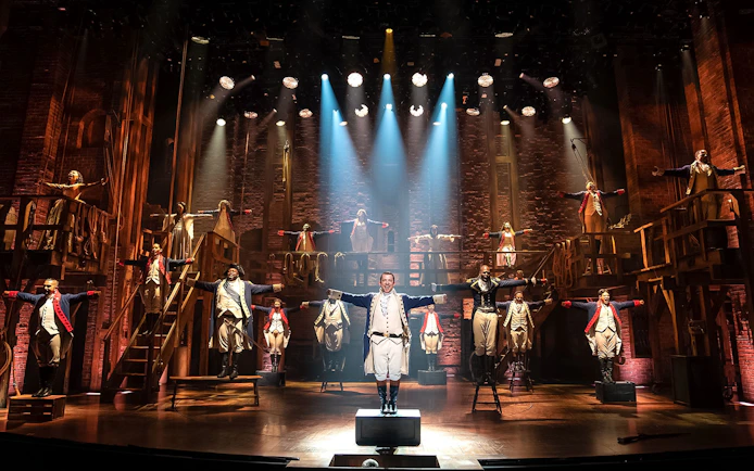 Stage performance of Hamilton cast in period costumes with dramatic lighting.