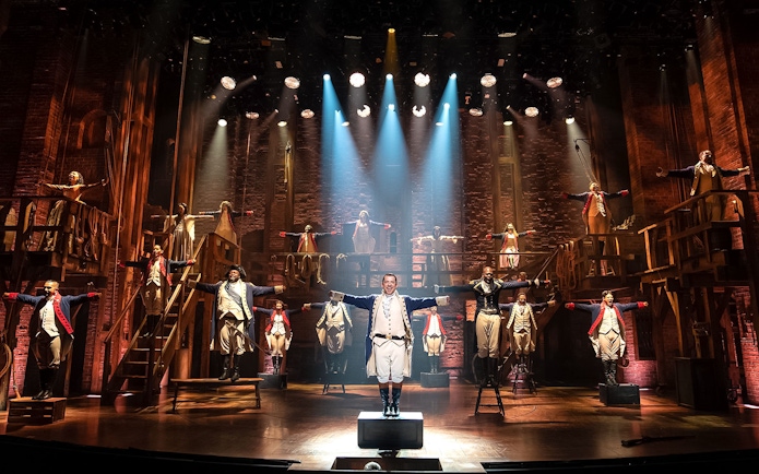 Stage performance of Hamilton cast in period costumes with dramatic lighting.
