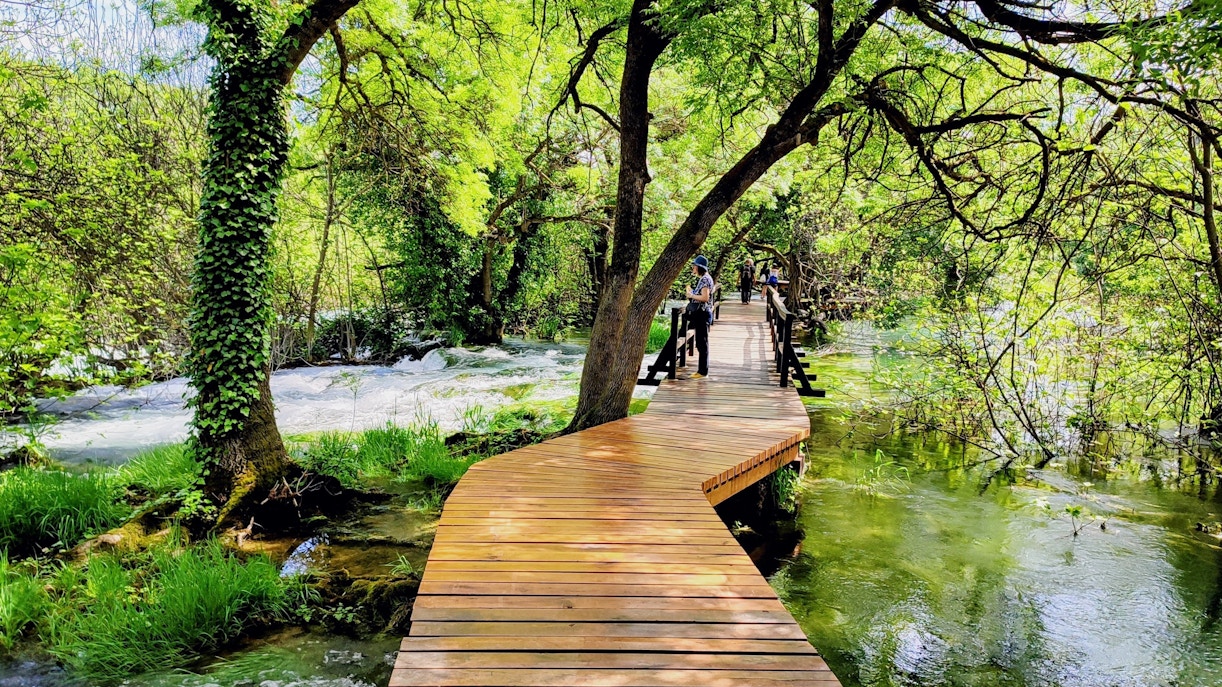 Wooden walkway through lush greenery and flowing water at Krka National Park.