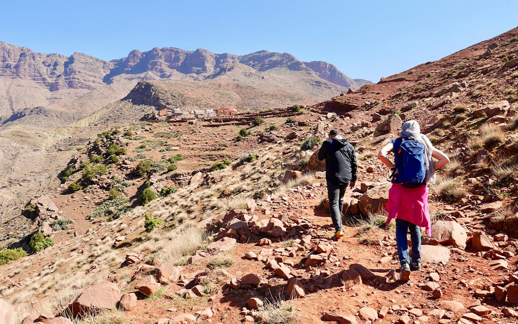 Hikers trekking rocky path in Ourika Valley towards traditional Berber village, Morocco.