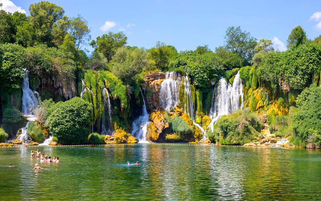Kravica Falls with tourists swimming in the emerald pool, surrounded by lush greenery.