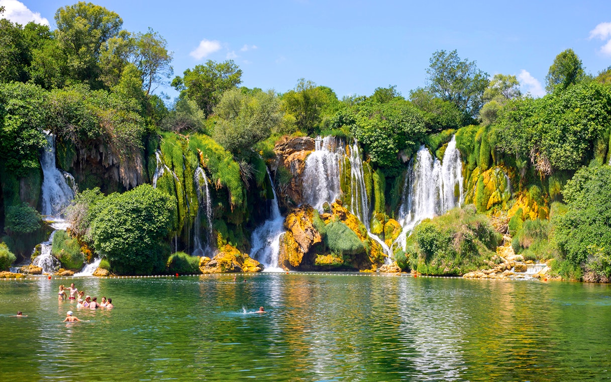 Kravica Falls with tourists swimming in the emerald pool, surrounded by lush greenery.