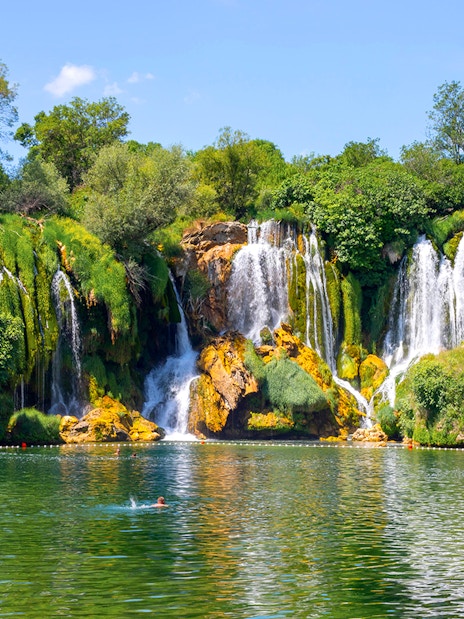Kravica Falls with tourists swimming in the emerald pool, surrounded by lush greenery.