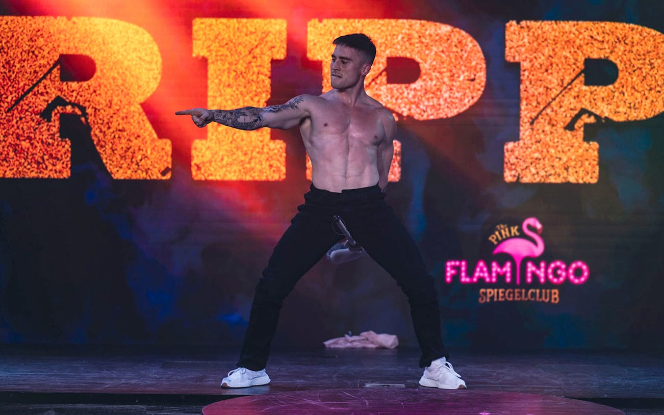 Performer on stage at The Pink Flamingo Ripped Show, striking a pose with vibrant backdrop.
