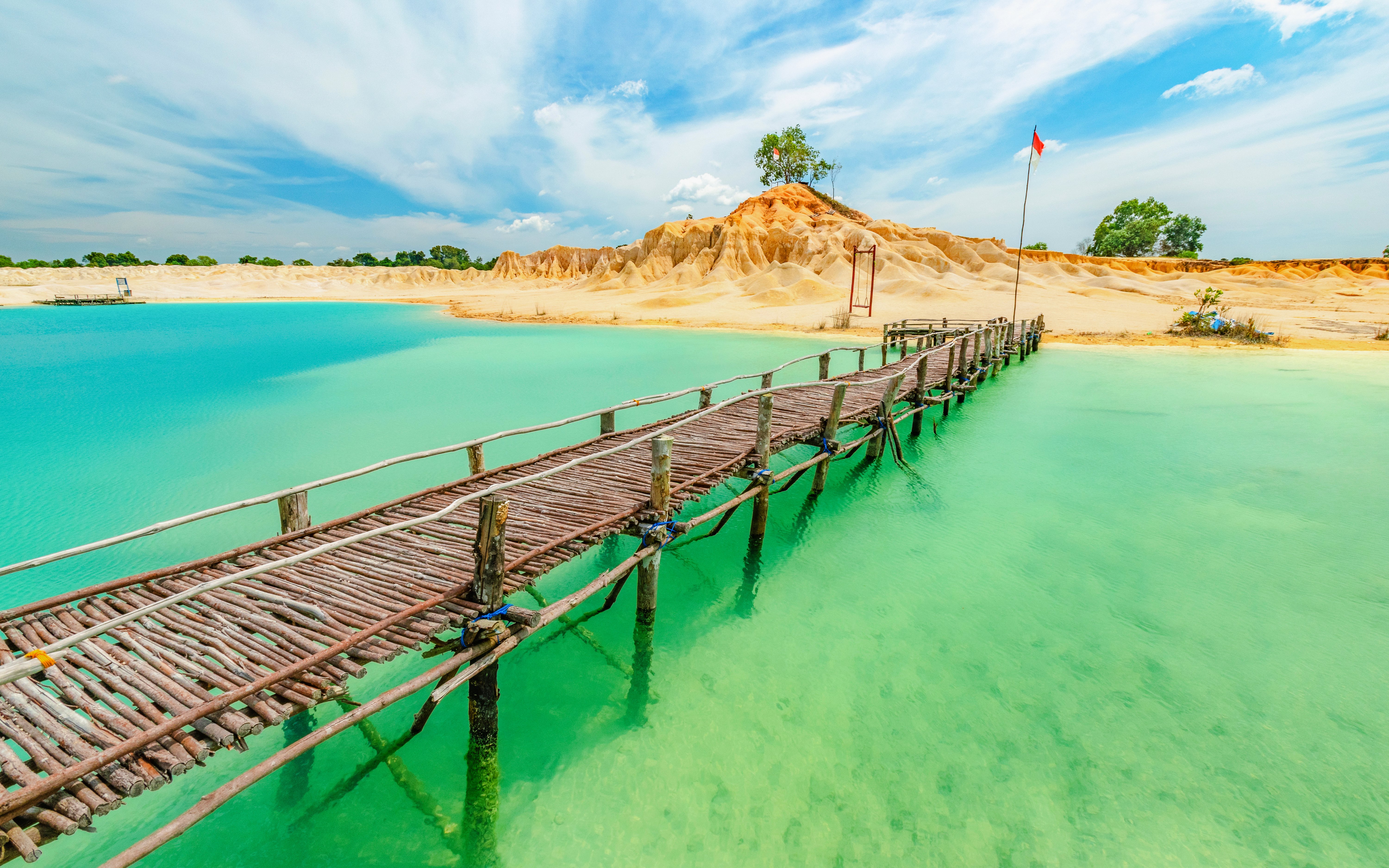Wooden bridge over turquoise water at Blue Lake, Bintan, with sandy hills in the background.