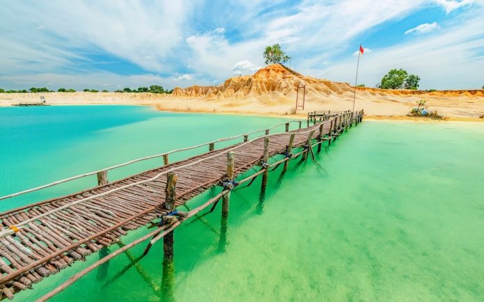 Wooden bridge over turquoise water at Blue Lake, Bintan, with sandy hills in the background.