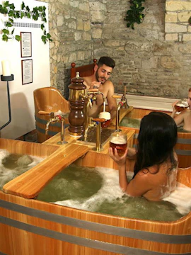 Guests enjoying a beer spa experience in wooden tubs with stone wall backdrop.