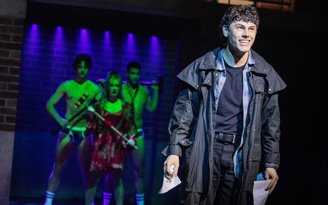 Performer on stage during Heathers: The Musical with actors in background.