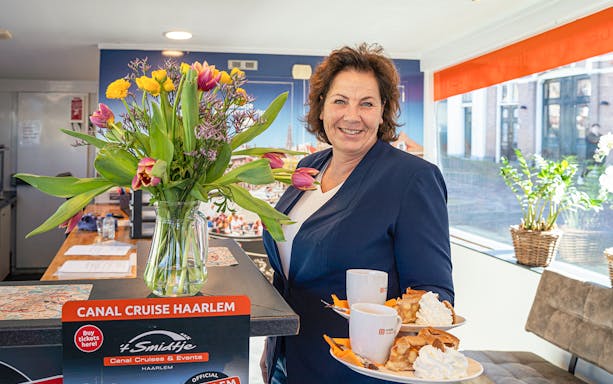 Woman serving coffee and pastries on a Canal Cruise Haarlem, with flowers and tour sign.