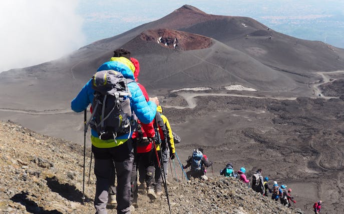 Hikers descending Mount Etna with summit crater in view, Sicily.