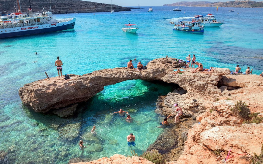 Tourists swimming and relaxing at the Blue Lagoon, Malta with boats in the background.