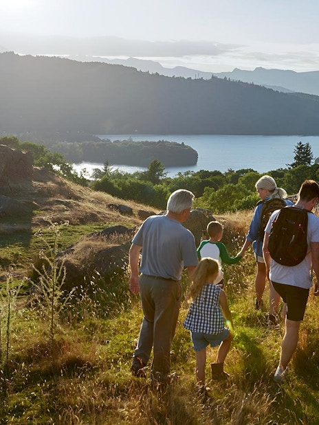 Group hiking in the Lake District with scenic lake and hills in the background.