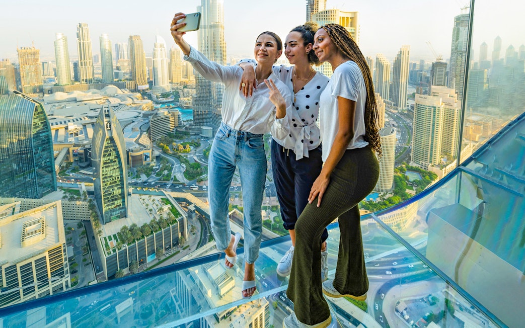 Three people taking a selfie on a glass observation deck overlooking a city skyline.
