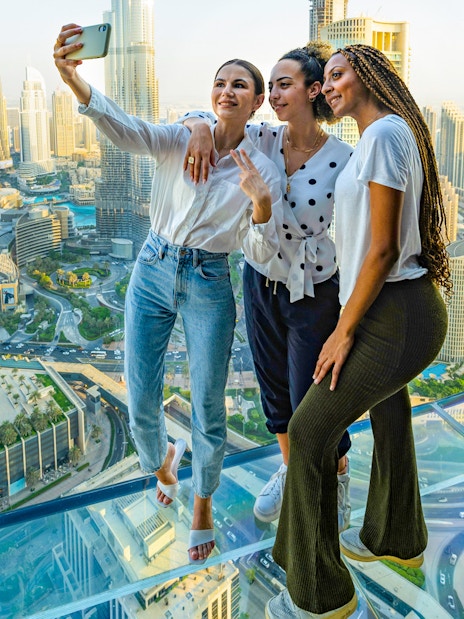 Three people taking a selfie on a glass observation deck overlooking a city skyline.