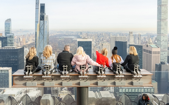Visitors seated on the beam at Top of the Rock Observation Deck, New York City skyline in view.