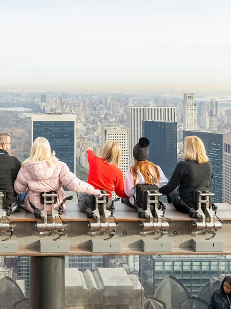 Visitors seated on the beam at Top of the Rock Observation Deck, New York City skyline in view.