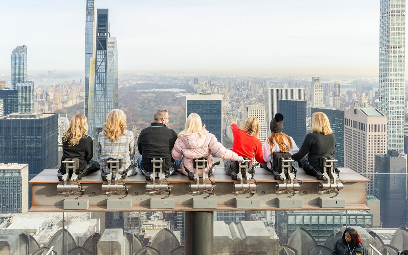 Visitors seated on the beam at Top of the Rock Observation Deck, New York City skyline in view.