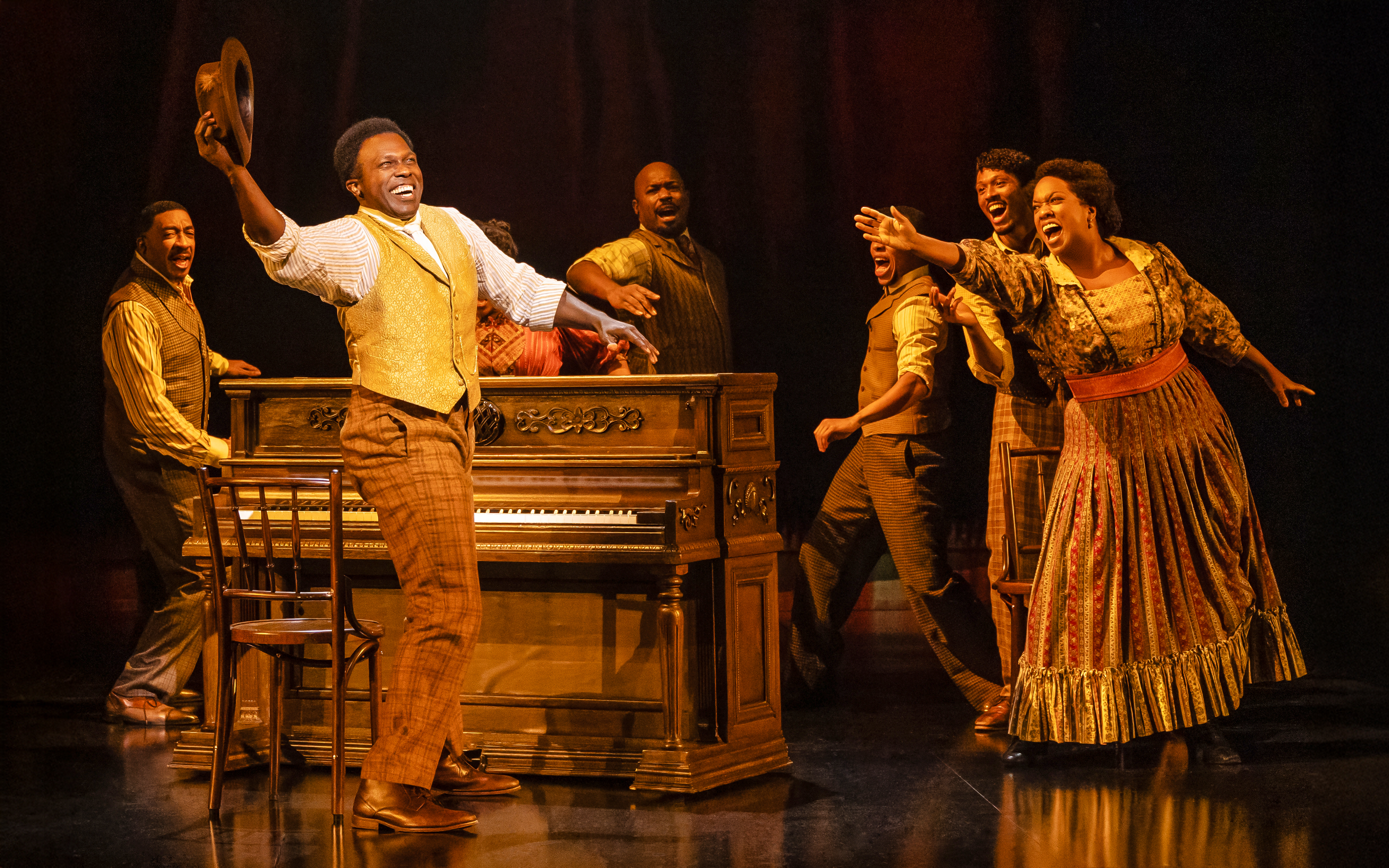 Performers singing around a piano in a scene from the musical Ragtime show.