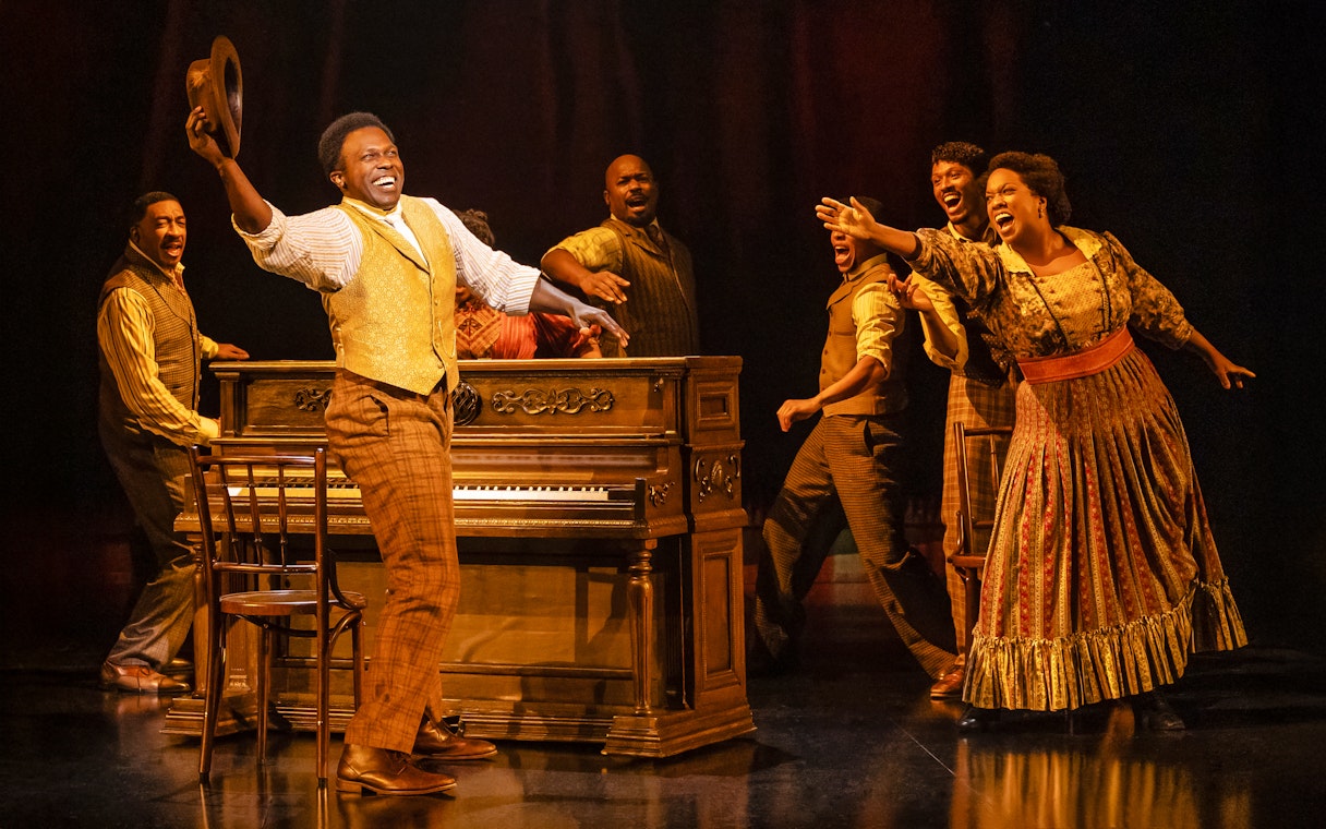 Performers singing around a piano in a scene from the musical Ragtime show.