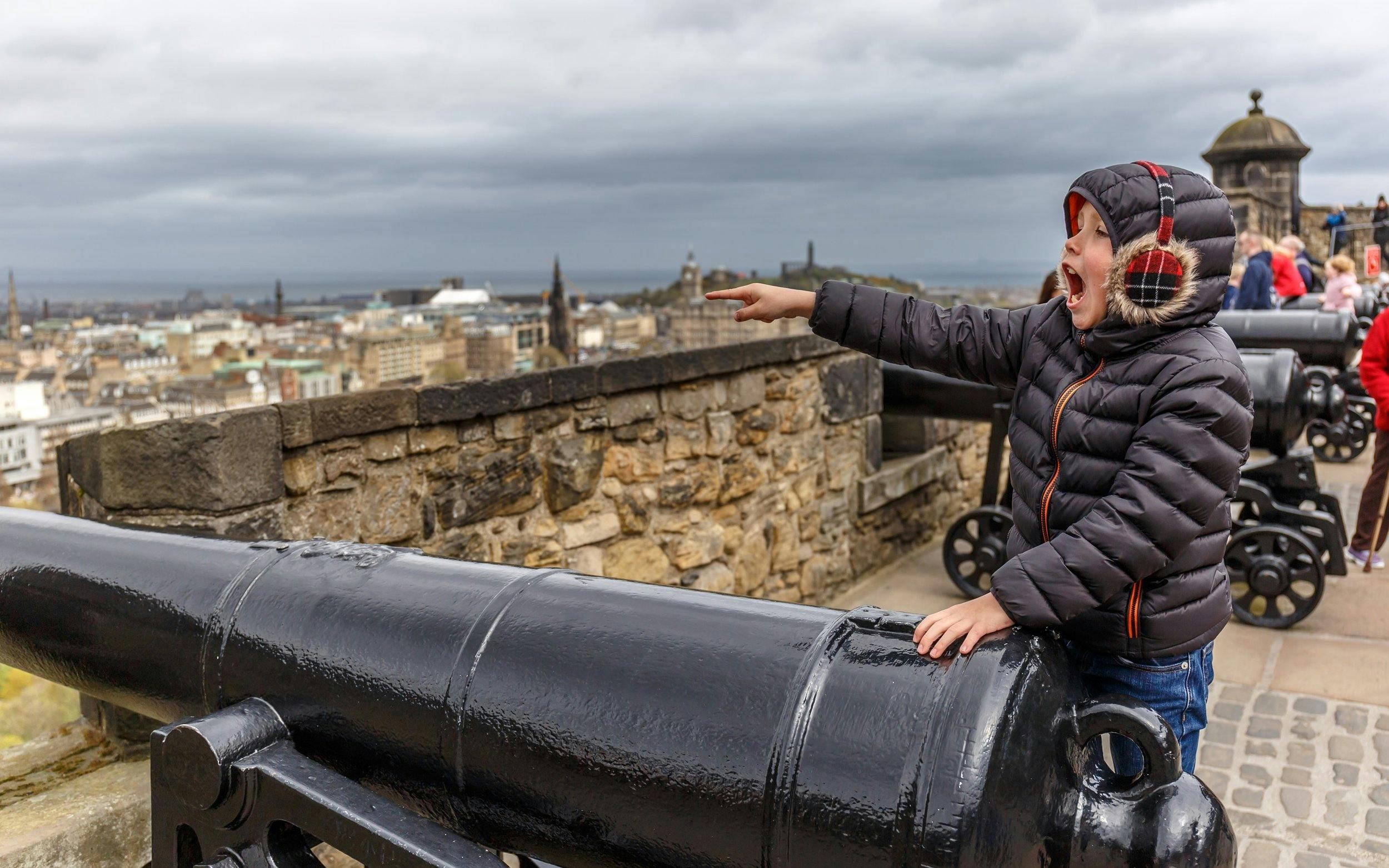Child pointing near cannons at Edinburgh Castle with city view in background.