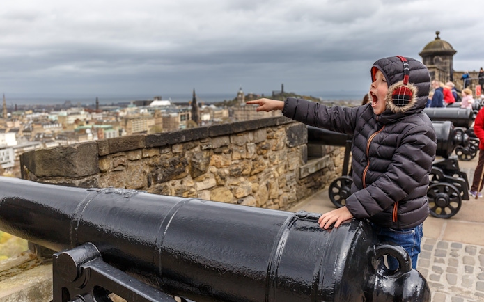 Child pointing near cannons at Edinburgh Castle with city view in background.