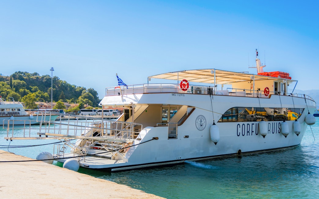 Corfu Cruises boat docked at a pier with scenic hillside in the background.