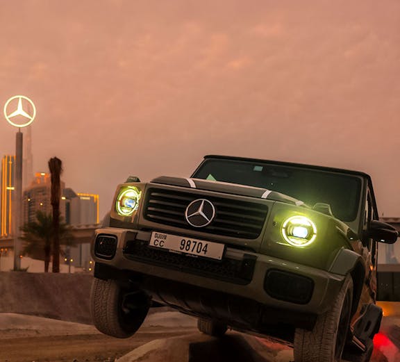 Mercedes-Benz SUV navigating terrain at sunset, Dubai skyline in background.