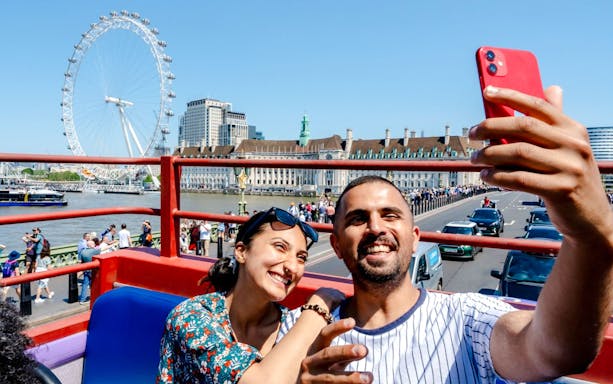 Tourists taking photos on a London hop-on hop-off bus with the London Eye in the background.