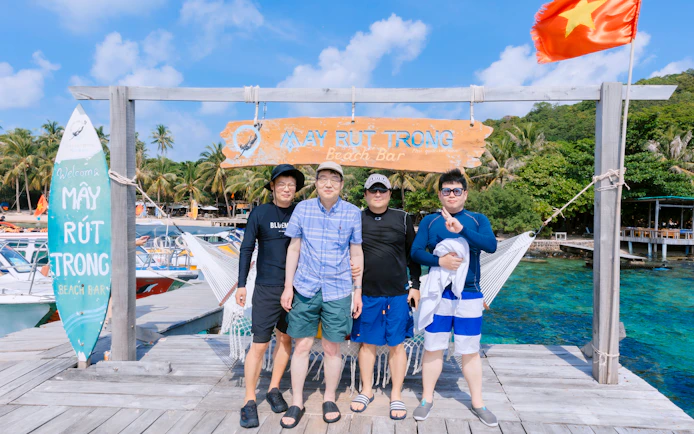 Group of tourists at May Rut Trong Beach Bar, Phu Quoc Island, with palm trees and boats in the background.