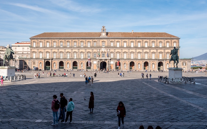 Royal Palace of Naples facade with equestrian statues and visitors in the foreground.