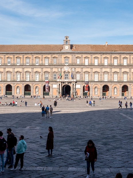 Royal Palace of Naples facade with equestrian statues and visitors in the foreground.