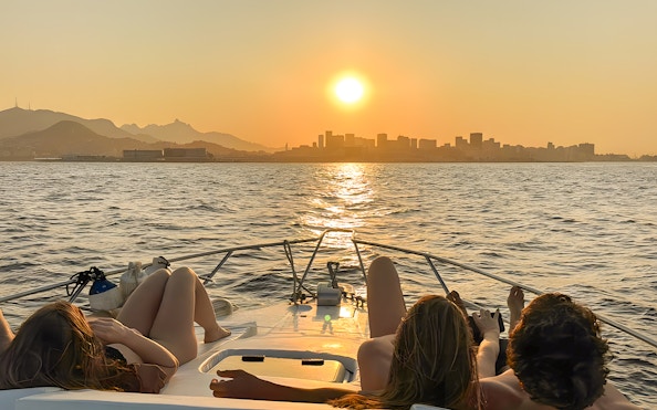 Tourists relaxing on a boat during a Rio de Janeiro sunset cruise with city skyline in view.