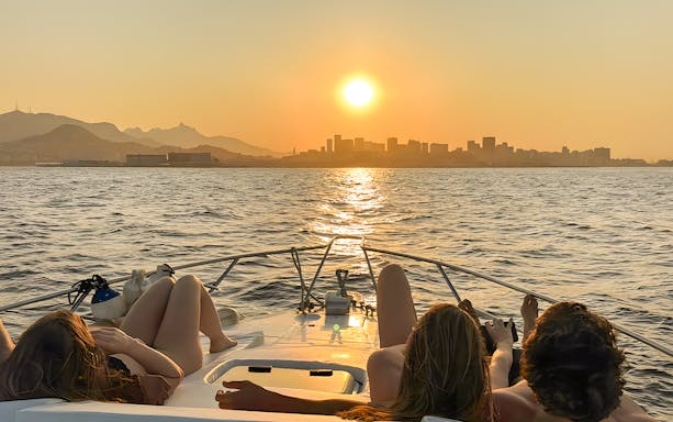 Tourists relaxing on a boat during a Rio de Janeiro sunset cruise with city skyline in view.