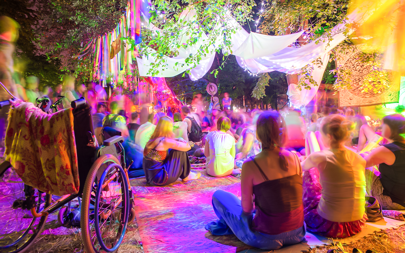 Outdoor gathering at Nuit Blanche with people sitting under colorful lights and decorations.